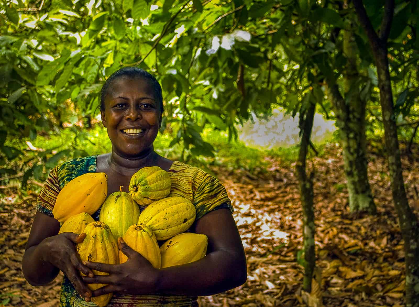 A woman stands smiling and holding a pile of bright yellow fruit surrounded by trees in the Amazon rainforest.