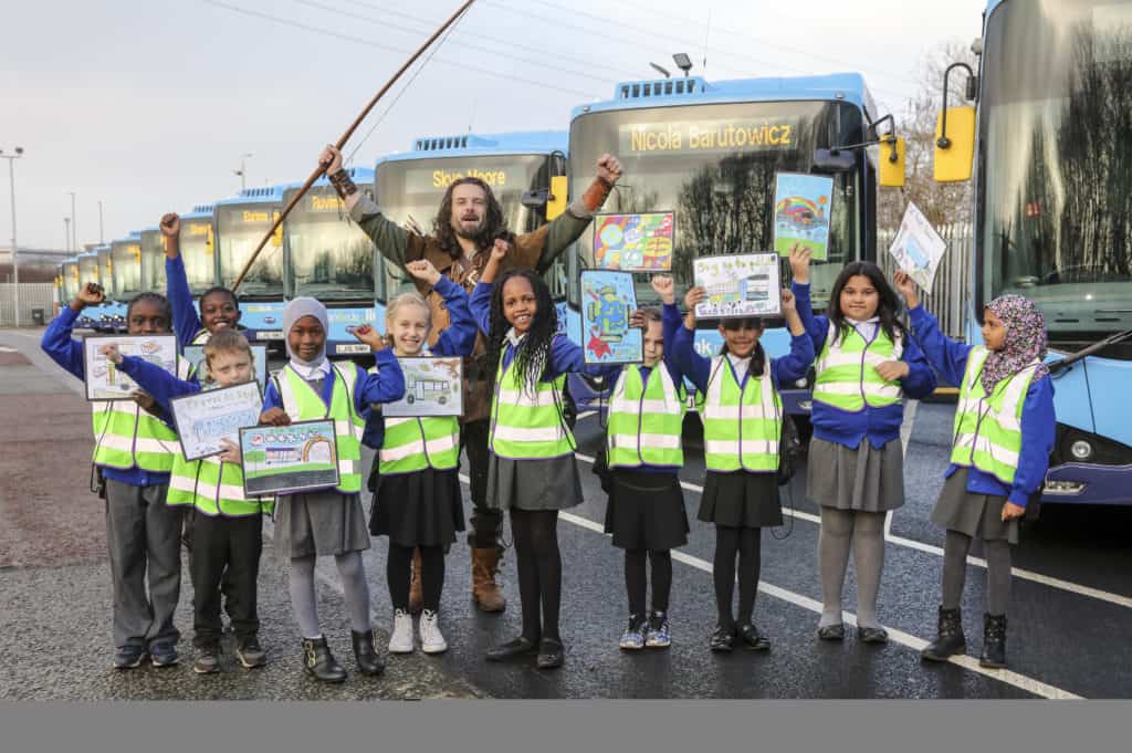 A group of children in hi-vis jackets holding drawings stand in front of new electric buses