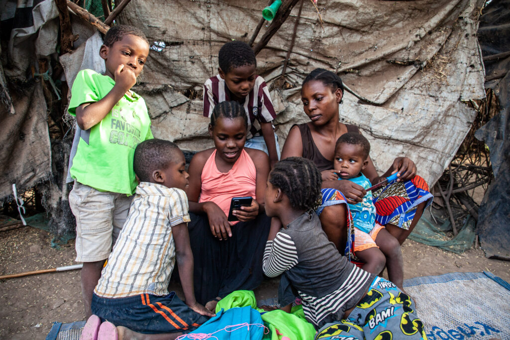 A mother and five young children in a refugee camp surround a young girl looking at a smart phone.