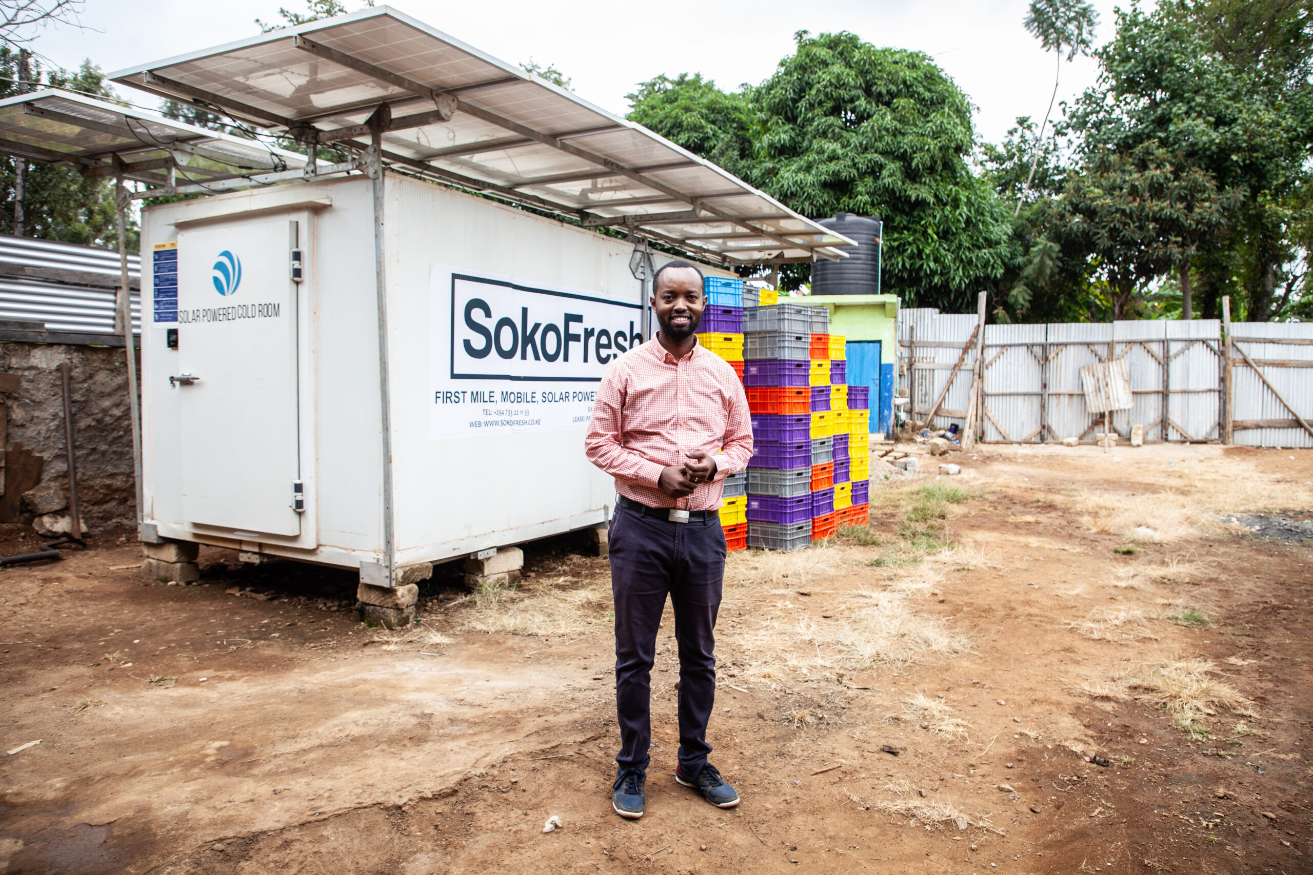 Photo of Dennis Karema, man in an orange collared shirt standing in front of stacked crates and a solar panel unit that reads "Soko Fresh"