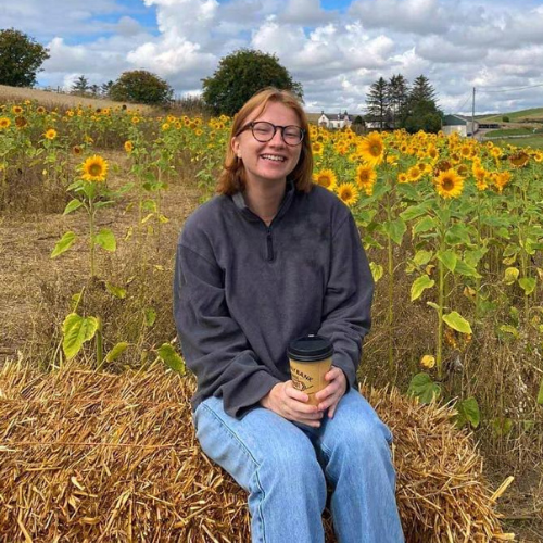 Ashden team member, Fran Tute sitting on haybail with a coffee and sunflowers in the background