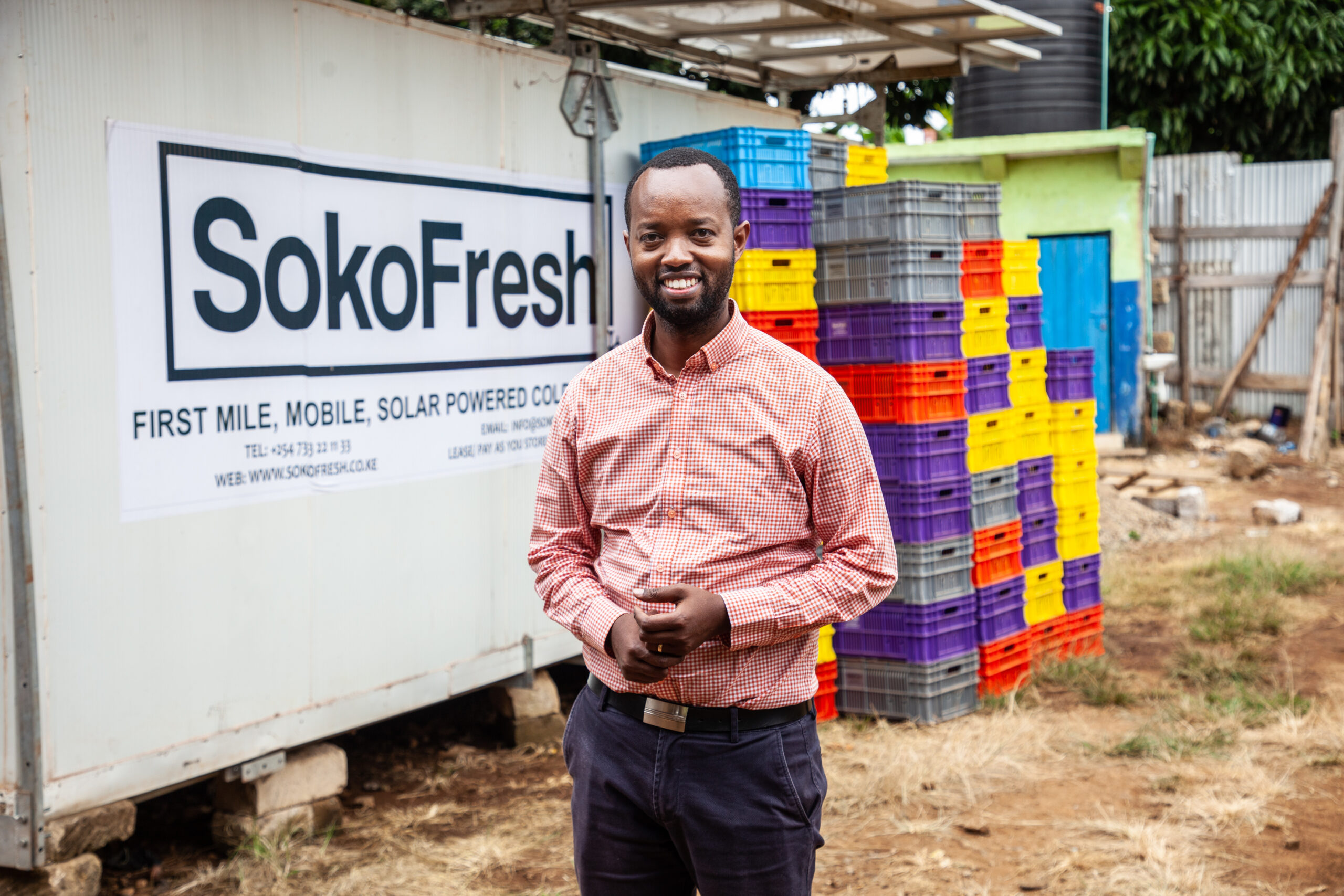 A man in an orange collared shirt stands in front of a stacked crates and a large unit that reads "Soko Fresh"
