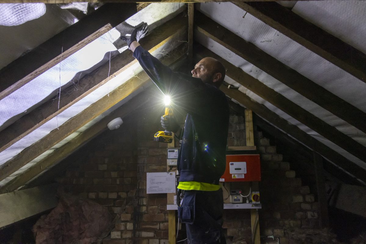 A man wearing work gloves holds a light and works on retrofitting an attic space.