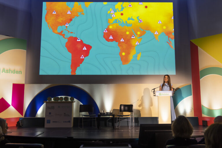 Woman speaking behind a podium on stage backed by a screen depicting a world map of the climate crisis