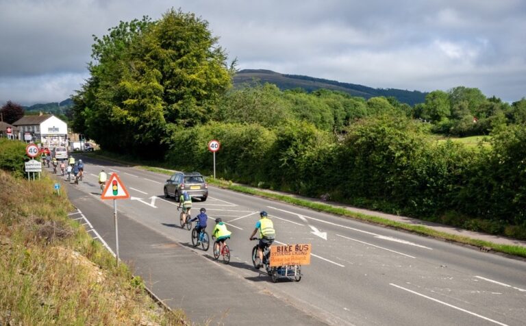 A group of students in high-vis vests cycle along a rural road. There is an adult beyond them on a bike with a sign that reads "Bike Bus. students cycling, please be patient"
