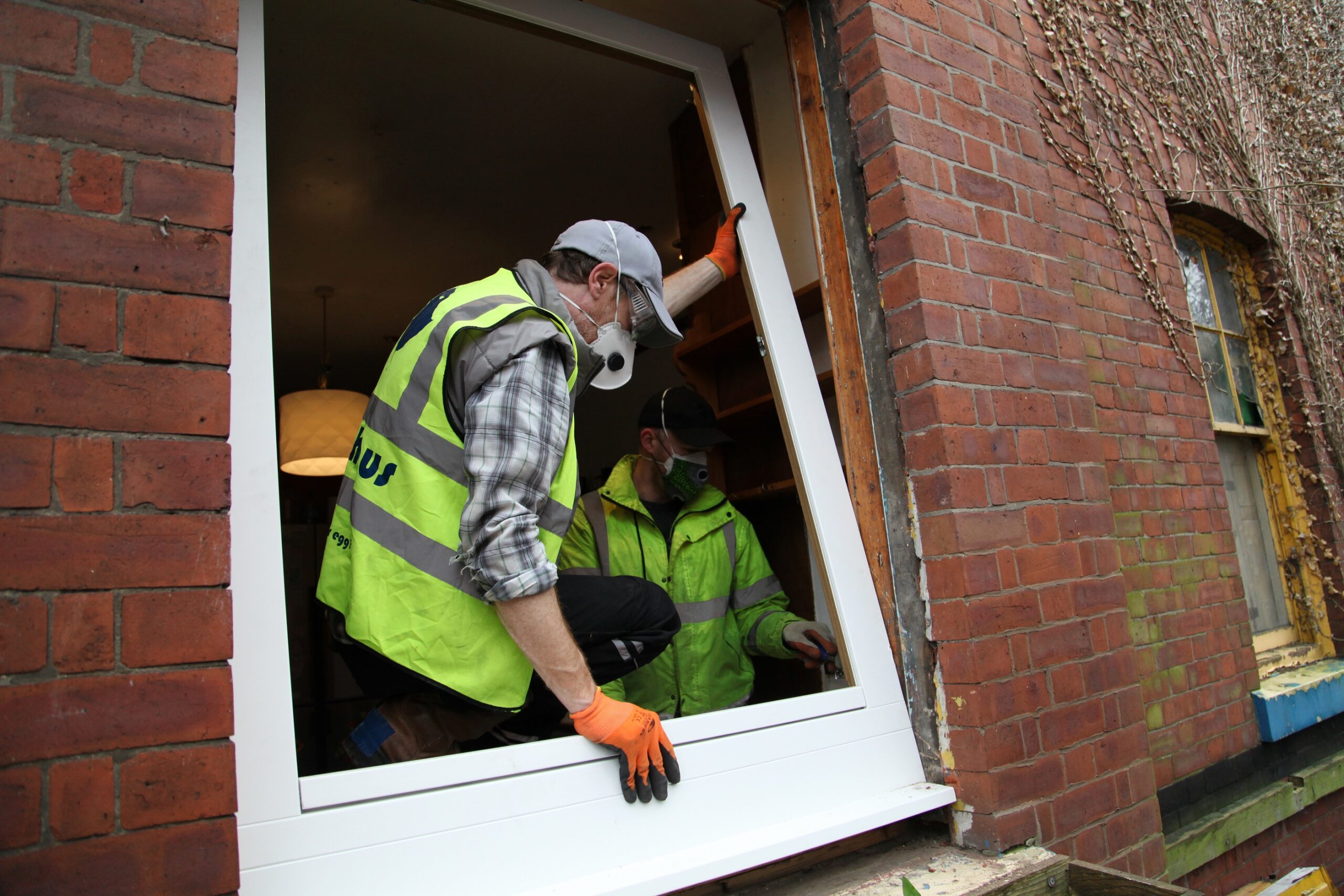 Two men in high-vis vests and work masks work on a window