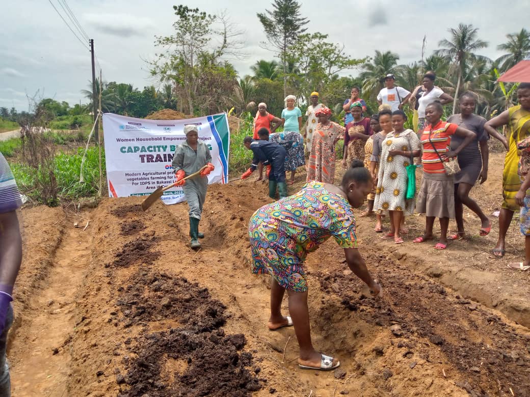 A woman with a shovel leads a training on irrigation while another woman assists by moving dirt. A group of 15 women observe.