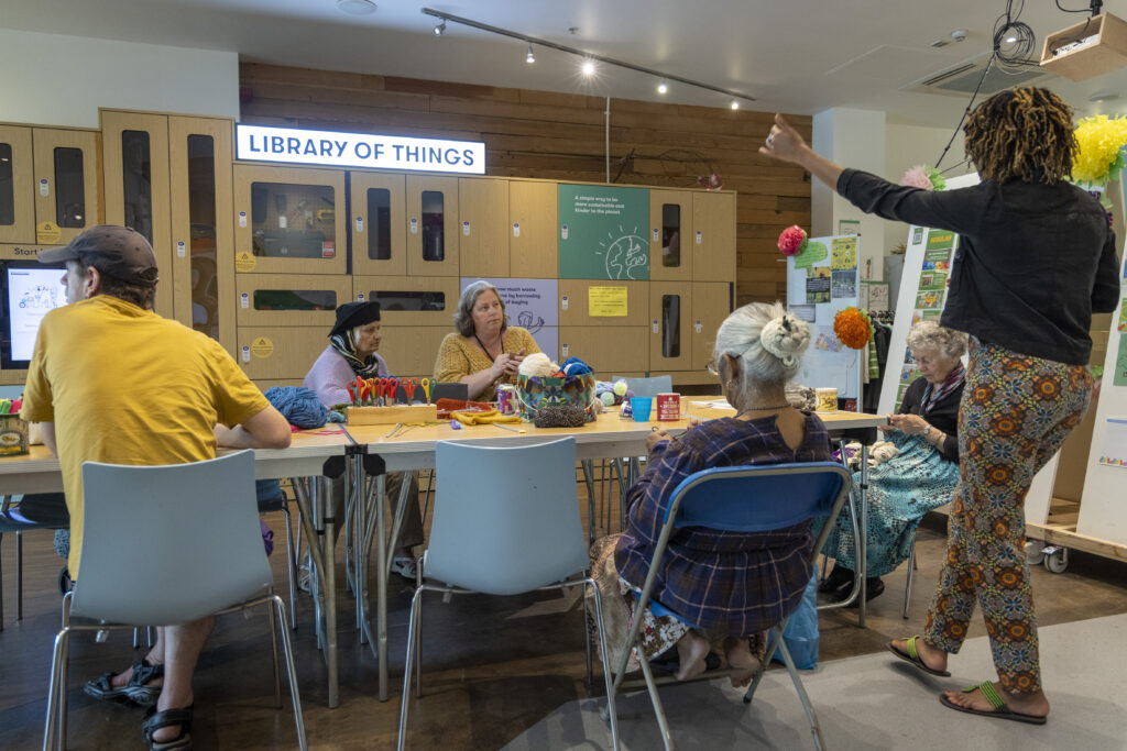 A group of people sitting at a table with a sign in the background saying 'library of things'