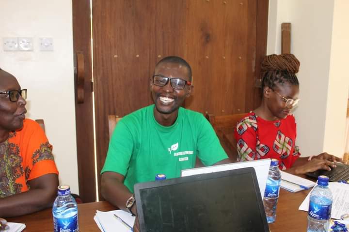 Three people sit at a table working on laptops. A man in a green shirt (middle) smiles big for the camera. 