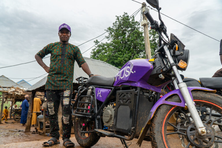 Man standing next to a purple motorcycle that reads "Husk" and wears a matching hat with the same logo.