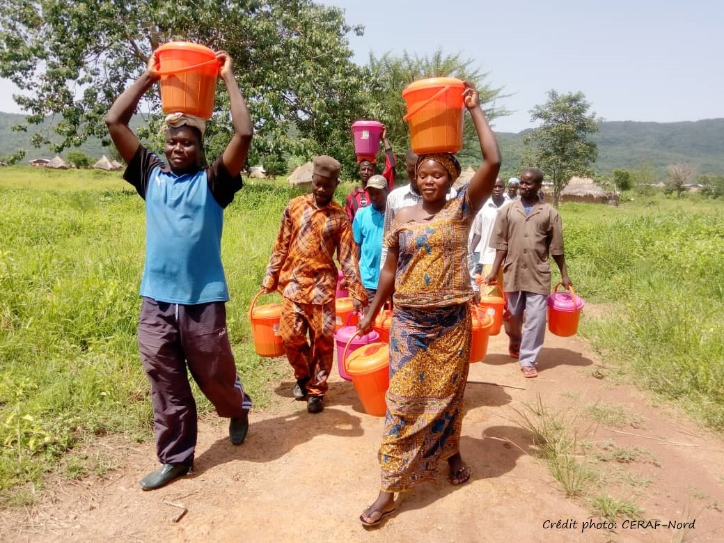 A group of people walking along a dirt road with buckets carrying water on their head