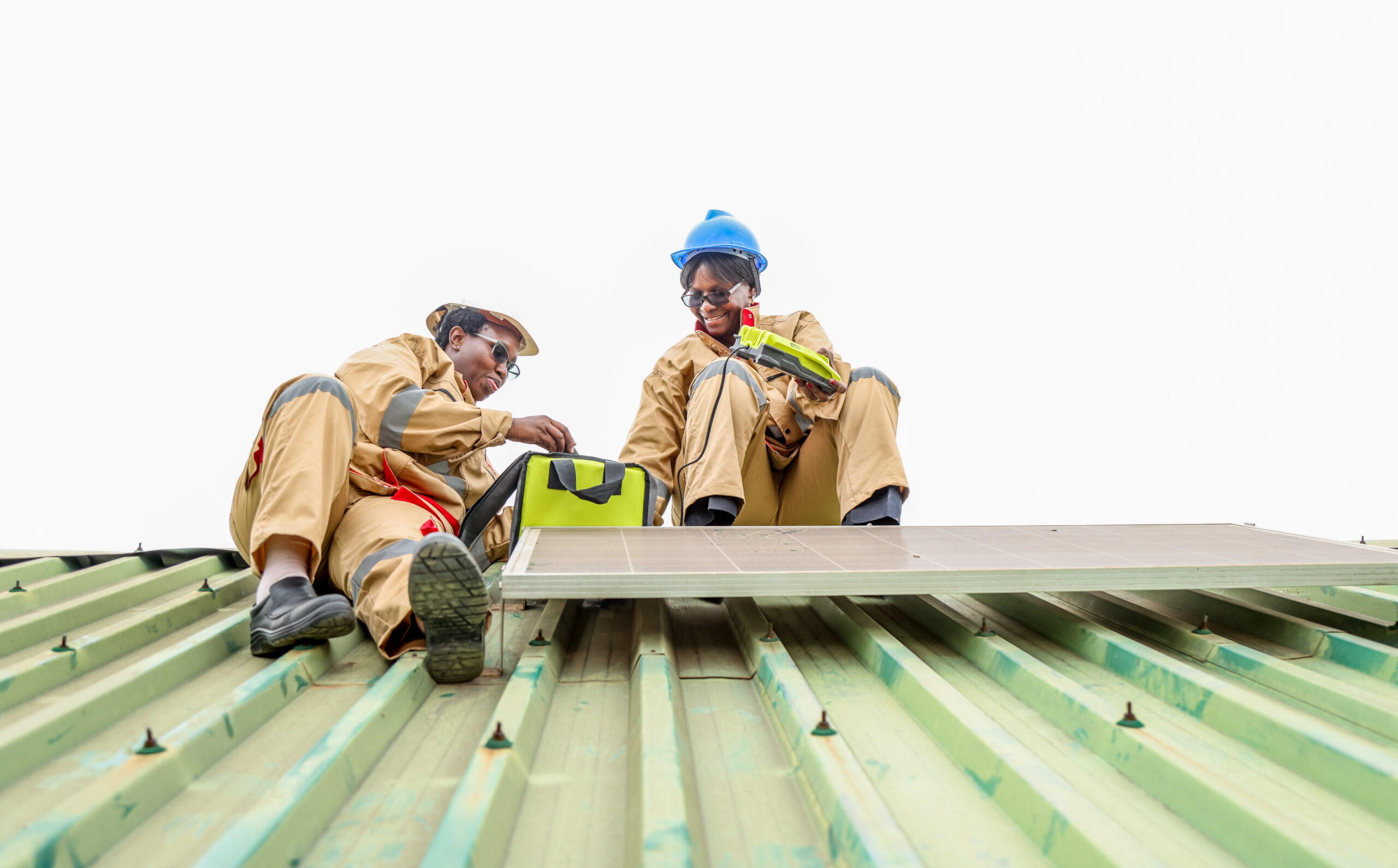 Two women in hard hats work on top of a metal roof installing a solar panel.