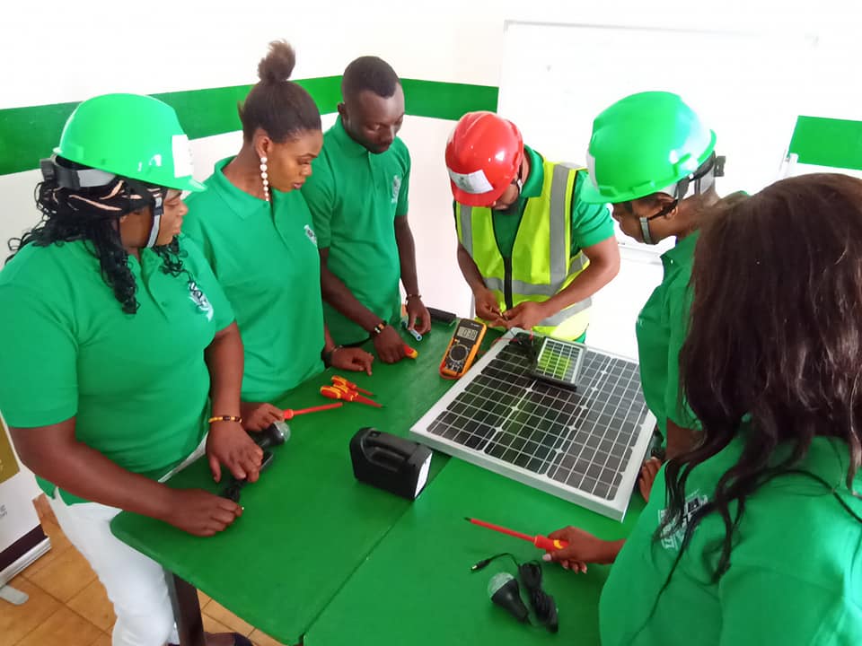 A man in a hard hat and high-vis vest repairs a solar lamp. 5 people in green polos stand around a table and observe his work.