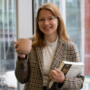 Woman with glasses smiling at the camera holding a pink mug and a book