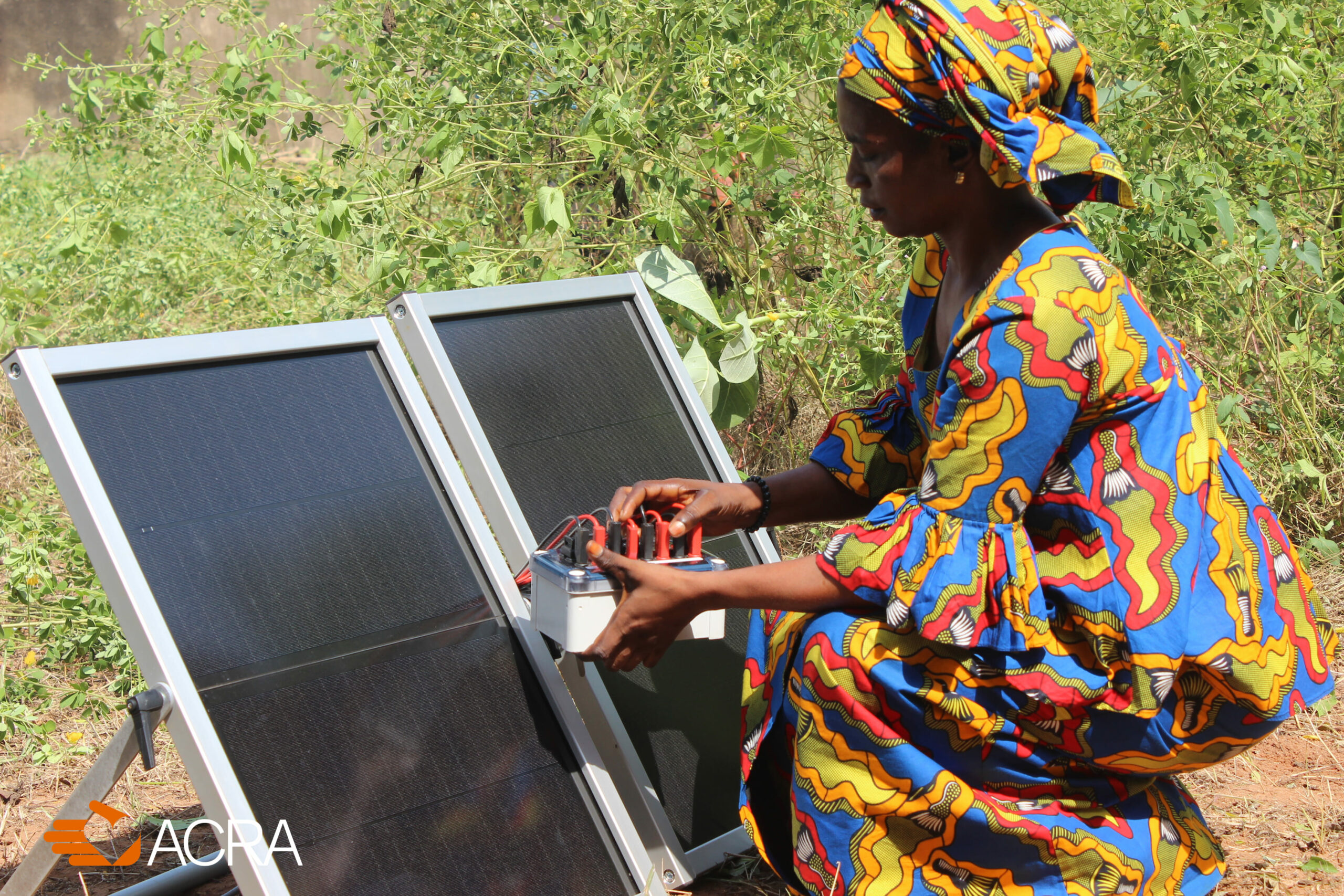 A woman kneels next to a solar panel and adjusts a box with electrical wiring.